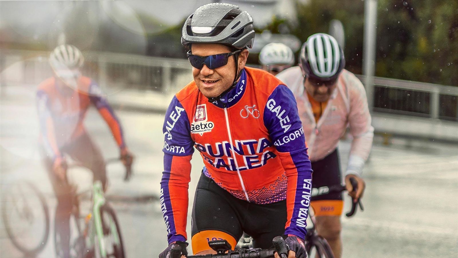 Cycliste en course portant un maillot et des lunettes de soleil, entouré d'autres coureurs sur route.