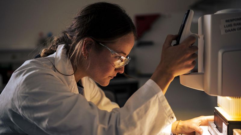 Layla Bral, scientifique chez Sanofi, se penchant sur un équipement de laboratoire, équipée d'une blouse et de lunettes de protection.